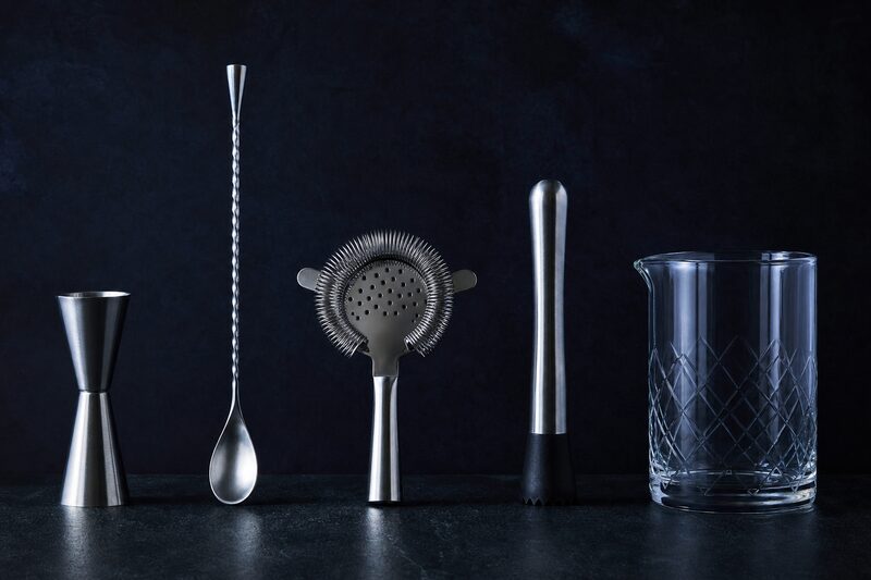 Attrezzatura da bar disposta in fila su un piano scuro: jigger doppio, cucchiaio da bar, strainer a molla, pestello e mixing glass.