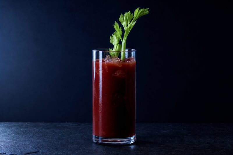 Bicchiere alto con un drink rosso denso e opaco a base di pomodoro su ghiaccio, un gambo di sedano infilato verticalmente.