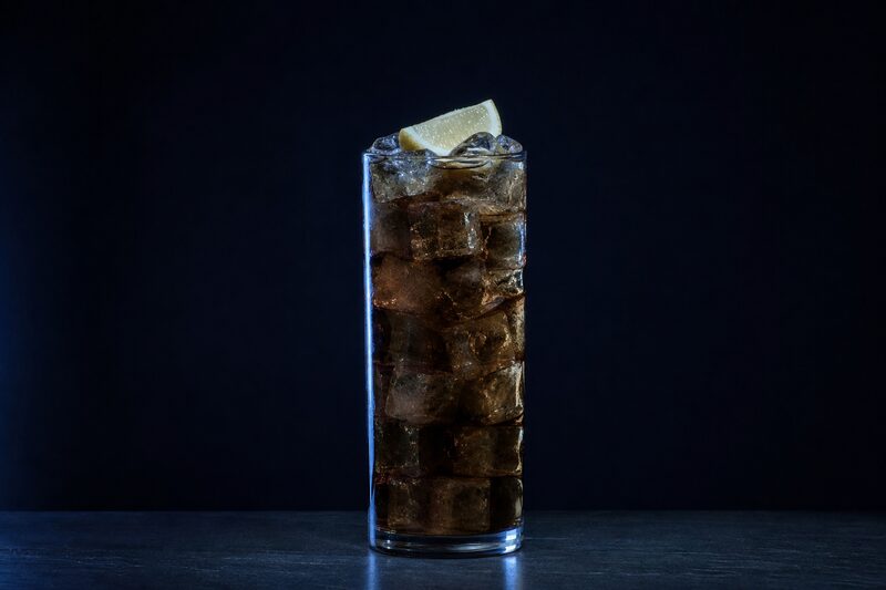 Bicchiere highball con una bevanda ghiacciata ambrata traslucida, cubetti impilati fino al bordo, uno spicchio di limone appoggiato sopra.