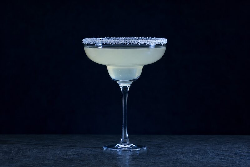 Calice da margarita con bordo salato regolare, riempito con un drink chiaro color paglia, i cristalli di sale nitidi nella luce fredda.