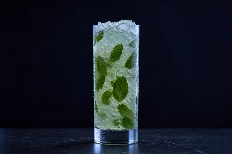 Bicchiere highball colmo di ghiaccio tritato con un drink chiaro dal riflesso verde, foglie di menta schiacciate contro la parete interna del vetro e condensa sulla superficie esterna.