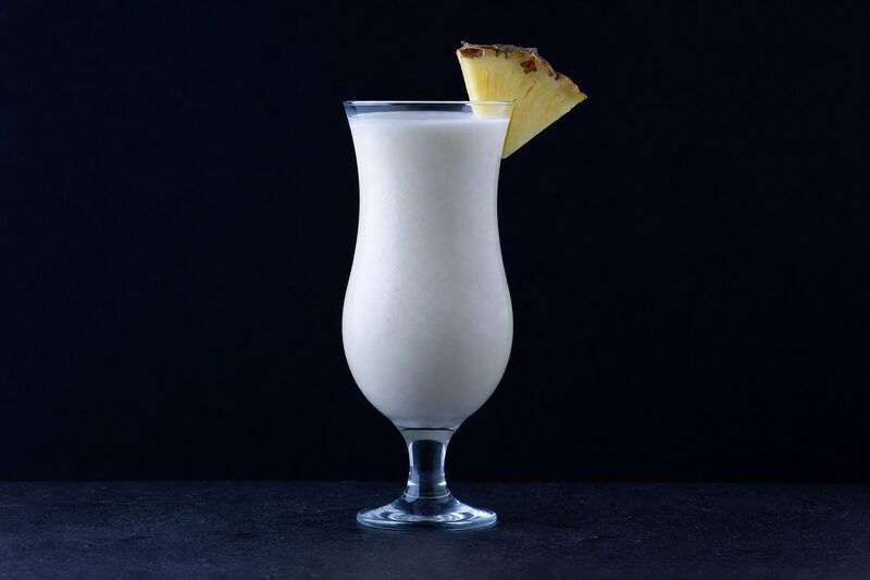 Bicchiere hurricane con una bevanda frullata densa e opaca, bianco crema, la superficie liscia e opaca, uno spicchio sottile di ananas sul bordo.